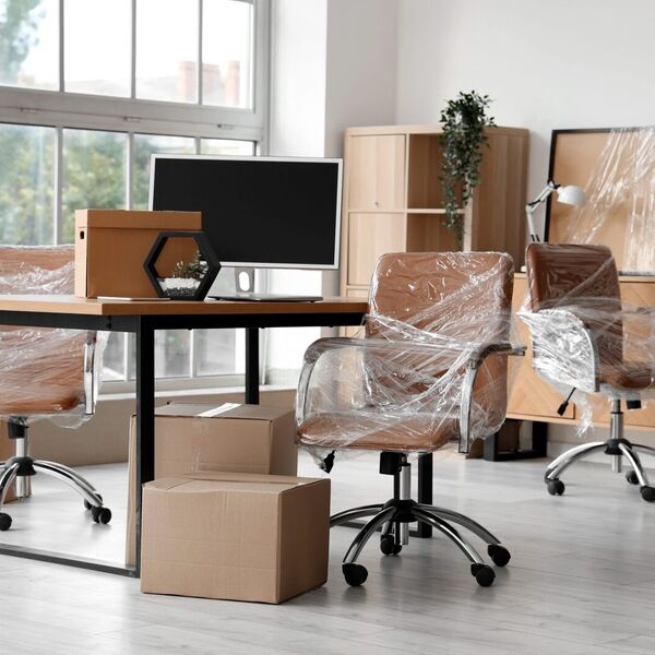 Office desk chairs wrapped in stretch film beside a desk, monitor and cardboard boxes ready for an office move