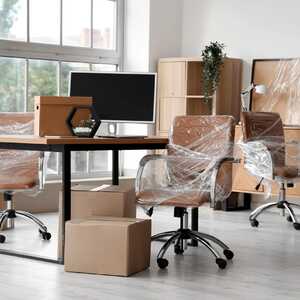 Office desk chairs wrapped in stretch film beside a desk, monitor and cardboard boxes ready for an office move