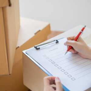 Hand ticking off a checklist on a clipboard resting on cardboard boxes while planning a move