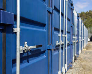 Row of blue shipping containers with metal locks and latches, set on a gravel path.