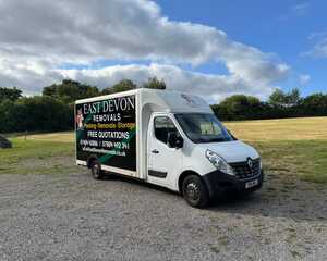 A white removal truck with "East Devon Removals" signage parked on gravel near grass.